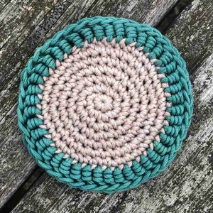 May include: A crocheted coaster with a teal border and a beige center. The coaster is made with a spiral pattern.