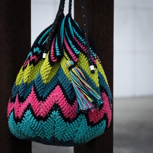 May include: A colorful crocheted bag with a zig-zag pattern in shades of pink, green, blue, and black. The bag has a drawstring closure and two long straps.