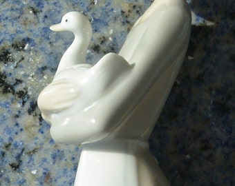 May include: A glossy porcelain figurine depicting a slender figure with light brown hair tied back, wearing a simple white dress. The figure is shown in profile, gently cradling a small white goose with a smooth, reflective finish and soft curves.