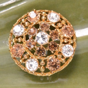 May include: A gold-toned brooch with a circular design featuring clear and brown rhinestones. The rhinestones are set in a delicate filigree pattern.