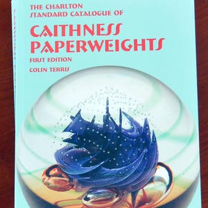 May include: A book titled "The Charlton Standard Catalogue of Caithness Paperweights, First Edition" by Colin Terris. The cover is light blue with red text and features a glass paperweight with a blue and gold design.