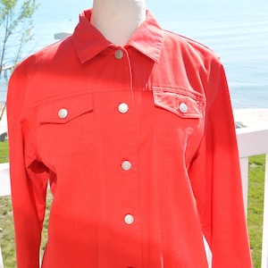 May include: A vibrant coral jacket with a button-down front and two chest pockets. The jacket has silver-coloured buttons and long sleeves. The garment is displayed on a mannequin, highlighting its tailored fit. The jacket is made of a smooth fabric, suitable for various occasions.