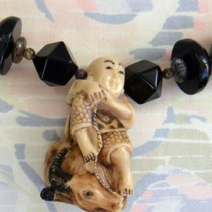 May include: A necklace made of black and brown beads with a carved ivory pendant of a man riding an ox. The man is holding a fish in his hands.