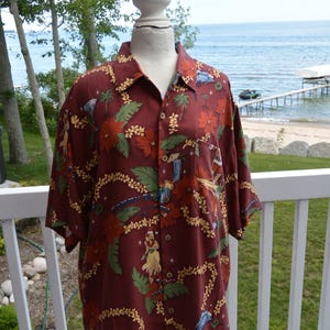 May include: Burgundy short-sleeve button-down shirt with a tropical floral and figure print. The shirt features a classic collar, a chest pocket, and a relaxed fit. The design includes palm trees, cocktails, and hula dancers.