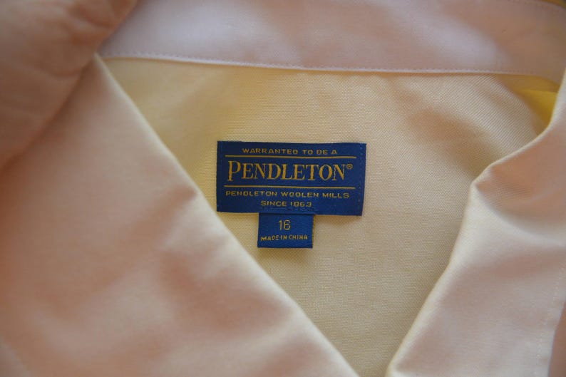 Puede incluir: Primer plano de una camisa amarillo claro con cuello blanco. Una etiqueta azul dice "WARRANTED TO BE A PENDLETON PENDLETON WOOLEN MILLS SINCE 1863". La etiqueta de talla dice "16 MADE IN CHINA".