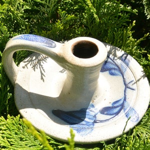 May include: A handmade ceramic candle holder with a handle and a wide base. The candle holder is off-white with blue floral designs and accents. The candle holder is sitting on a bed of green foliage.