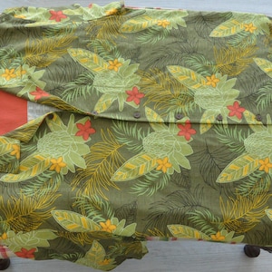 K&ouml;nnte beinhalten: Olivgr&uuml;nes Hemd mit tropischem Blumenmuster mit gelben, gr&uuml;nen und roten Bl&uuml;ten und Bl&auml;ttern. Das Hemd ist auf einem gemusterten Kissen gefaltet. Das Hemd hat ein Etikett.