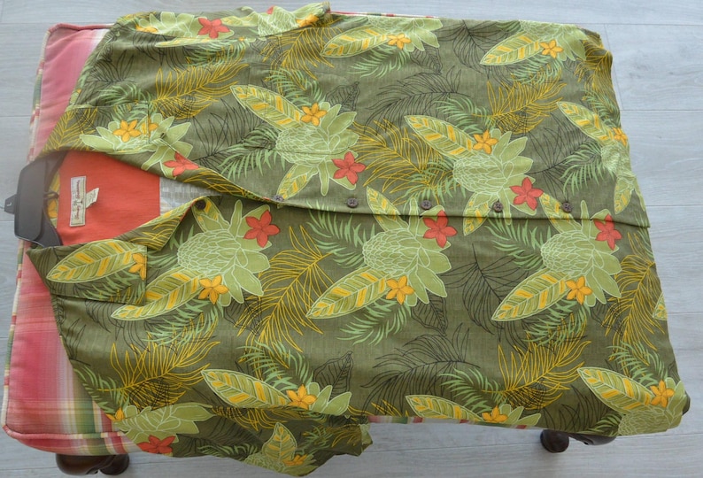 K&ouml;nnte beinhalten: Olivgr&uuml;nes Hemd mit tropischem Blumenmuster mit gelben, gr&uuml;nen und roten Bl&uuml;ten und Bl&auml;ttern. Das Hemd ist auf einem gemusterten Kissen gefaltet. Das Hemd hat ein Etikett.