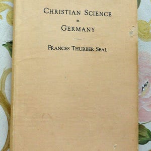 May include: A vintage book titled "Christian Science in Germany" by Frances Thurber Seal. The book has a light tan cover with black text. The cover shows signs of wear and tear.