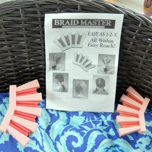 May include: A "Braid Master" instruction sheet with illustrations of hairstyles, accompanied by two pink and coral-colored hair braiding tools. The sheet reads "Easy as 1-2-3! All Within Easy Reach!" on a blue floral patterned fabric.