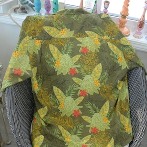 K&ouml;nnte beinhalten: Olivgr&uuml;nes Hemd mit tropischem Blumenmuster. Das Design zeigt gro&szlig;e stilisierte Blumen in Gr&uuml;n-, Gelb- und Rott&ouml;nen vor einem Hintergrund aus Palmfarnen. Das Hemd wird auf einem Korbstuhl pr&auml;sentiert.