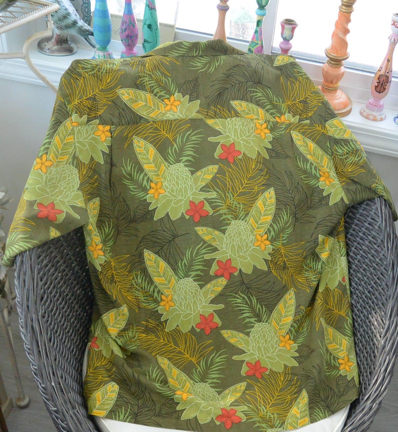 K&ouml;nnte beinhalten: Olivgr&uuml;nes Hemd mit tropischem Blumenmuster. Das Design zeigt gro&szlig;e stilisierte Blumen in Gr&uuml;n-, Gelb- und Rott&ouml;nen vor einem Hintergrund aus Palmfarnen. Das Hemd wird auf einem Korbstuhl pr&auml;sentiert.