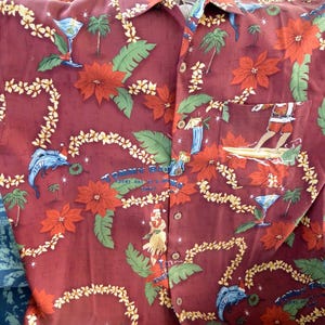 May include: Burgundy button-down shirt with a tropical print. The design features red poinsettias, palm trees, and surfers. The shirt has a pocket and a collar. The text "TOMMY BAHAMA EVERY DAY IS A HOLIDAY" is visible.