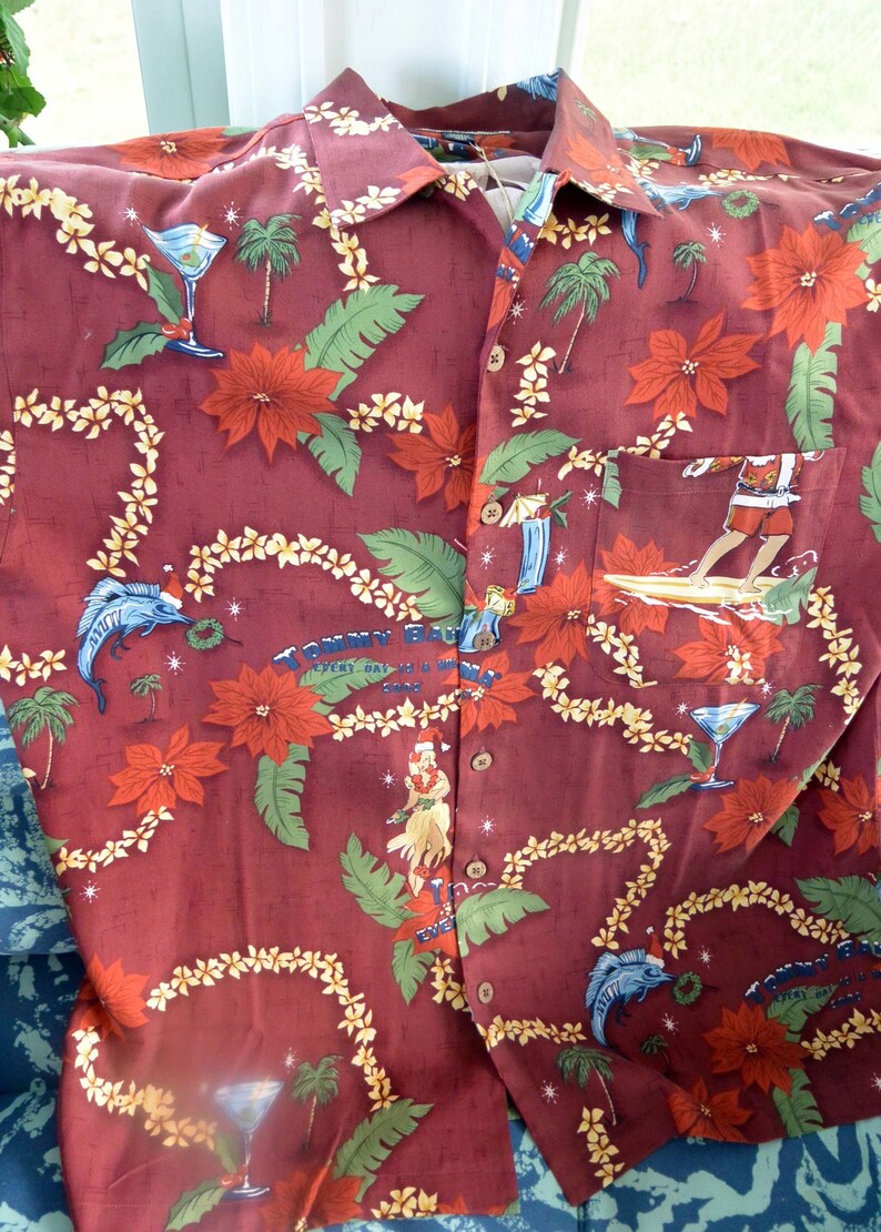 May include: Burgundy button-down shirt with a tropical print. The design features red poinsettias, palm trees, and surfers. The shirt has a pocket and a collar. The text "TOMMY BAHAMA EVERY DAY IS A HOLIDAY" is visible.