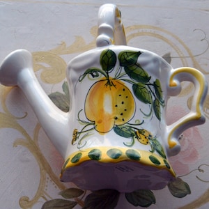 May include: White ceramic watering can with a yellow lemon design and green leaves. The watering can has a handle and a spout.