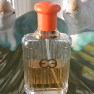 May include: A clear glass perfume bottle with an orange cap and gold accents. The bottle contains a light amber liquid and features a logo with two intertwined letter Es. The words "Eau de toilette" are printed at the bottom.