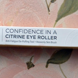 Op de afbeelding: Een witte rechthoekige doos met de tekst "CONFIDENCE IN A CITRINE EYE ROLLER" en het merklogo "it". De doos zegt ook "Anti-Fatigue De-Puffing Tool + Heavenly Skin Brush". De doos staat op een bloemenachtergrond.