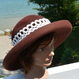 Puede incluir: Un sombrero de fieltro marrón con ala ancha y un ribete trenzado blanco. El sombrero está sentado en una cabeza de maniquí.