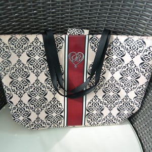 May include: A beige tote bag featuring a black damask pattern. The bag has a central vertical red stripe with a silver heart-shaped emblem. Black straps are attached to the top of the bag.