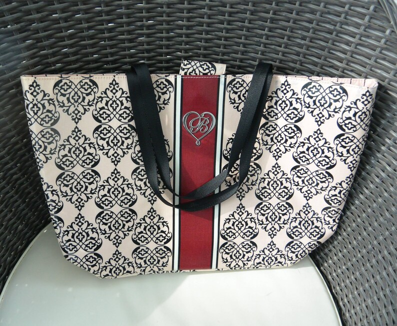 May include: A beige tote bag featuring a black damask pattern. The bag has a central vertical red stripe with a silver heart-shaped emblem. Black straps are attached to the top of the bag.