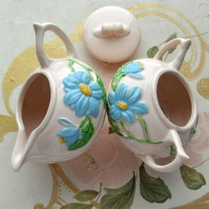 Peut inclure: Un ensemble vintage de pot à crème et sucrier rose avec des motifs floraux bleus et verts. Le pot à crème et le sucrier ont une forme arrondie et un motif floral délicat.