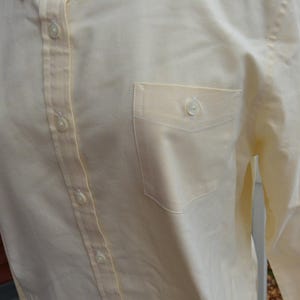 Puede incluir: Camisa amarilla p&aacute;lida con botones y bolsillo en el pecho. La camisa tiene mangas largas y cuello cl&aacute;sico. Los botones son peque&ntilde;os y blancos. La tela parece ser una mezcla ligera de algod&oacute;n o lino.