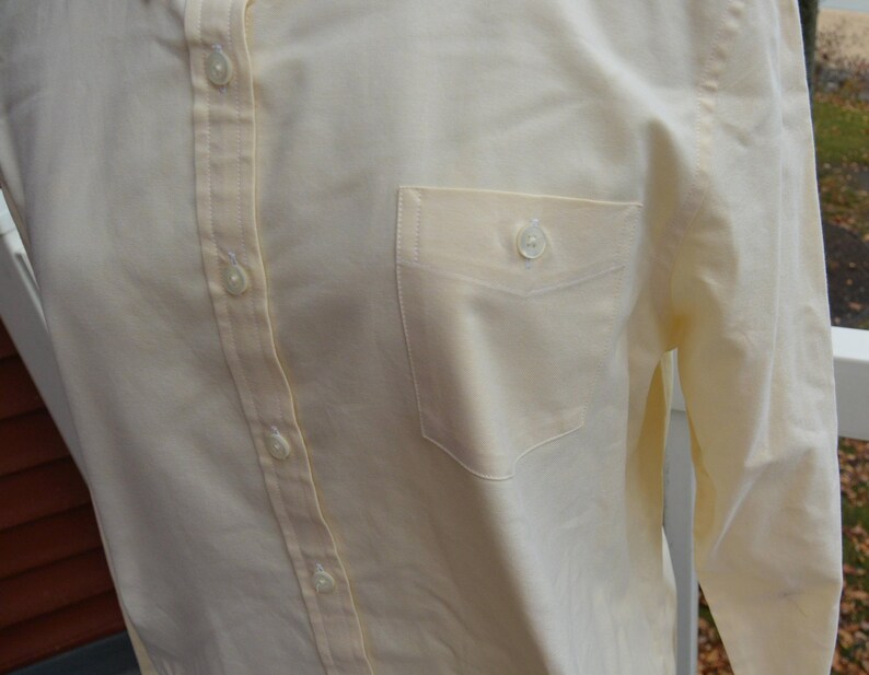 Puede incluir: Camisa amarilla p&aacute;lida con botones y bolsillo en el pecho. La camisa tiene mangas largas y cuello cl&aacute;sico. Los botones son peque&ntilde;os y blancos. La tela parece ser una mezcla ligera de algod&oacute;n o lino.