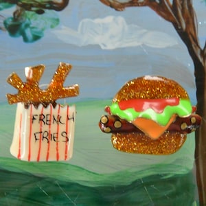 May include: A pair of glittery food-themed pins. One pin is a red and white french fry container with the words "French Fries" on it. The other pin is a glittery brown hamburger with green lettuce, red tomato, and yellow cheese.