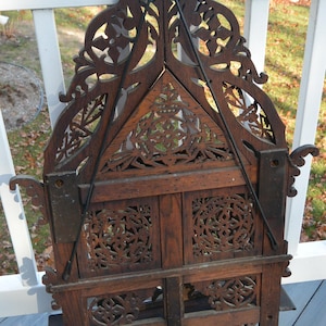 May include: A dark brown wooden wall hanging with intricate carved designs. The hanging is shaped like a triangle with a pointed top and a curved bottom. It has a metal hook on the top for hanging.