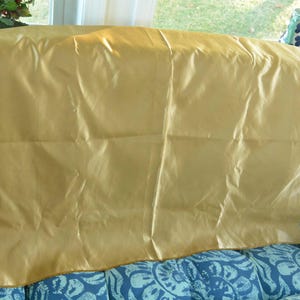 May include: A golden-yellow, rectangular fabric draped over a blue patterned surface. The fabric has a shiny, slightly wrinkled appearance, suggesting a satin or silk material. The edges are neatly finished, and the fabric is the primary focus of the image.