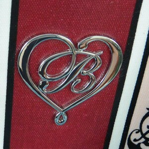 May include: A silver heart-shaped emblem with the letter "B" at its centre, set against a textured, deep red backdrop. The design is framed by black and white stripes and a floral pattern. The emblem has a polished, reflective surface.
