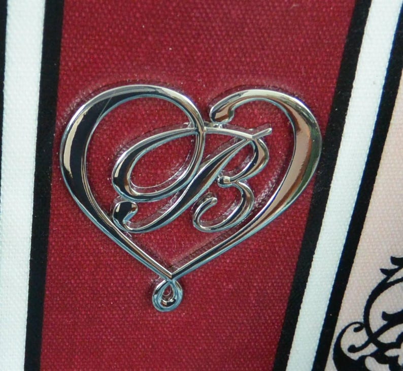 May include: A silver heart-shaped emblem with the letter "B" at its centre, set against a textured, deep red backdrop. The design is framed by black and white stripes and a floral pattern. The emblem has a polished, reflective surface.