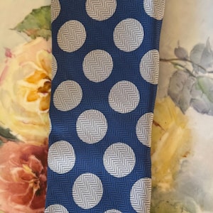 May include: A navy blue tie with a repeating pattern of large white circles filled with a herringbone design. A small gold emblem is near the top. The tie is folded on a decorative tray.