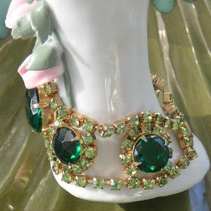 May include: A gold-tone bracelet featuring large, round, dark emerald green faceted stones surrounded by smaller, light lime green rhinestones. The bracelet is constructed with a metal link chain setting that holds the stones in a circular pattern.