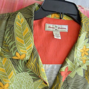 K&ouml;nnte beinhalten: Ein Hemd mit tropischem Blumendruck in Gr&uuml;n-, Gelb- und Oranget&ouml;nen. Das Hemd hat einen Kragen und ein leuchtend orangefarbenes Innenfutter. Das Etikett lautet "Tommy Bahama."