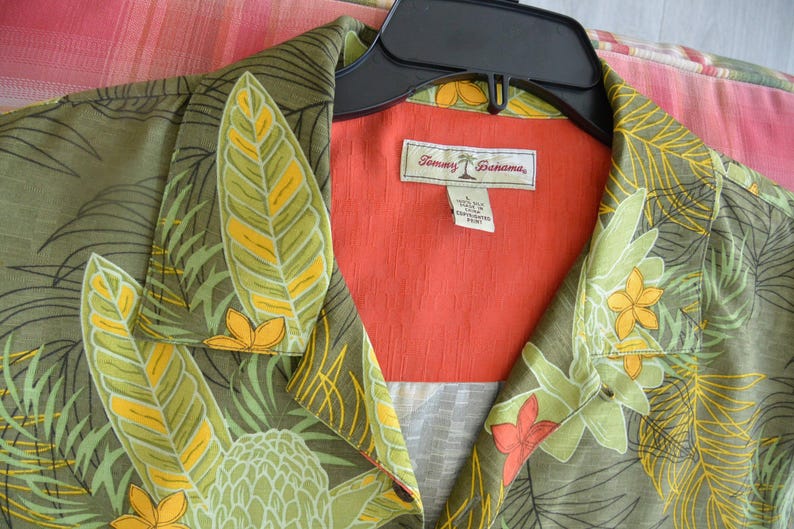 K&ouml;nnte beinhalten: Ein Hemd mit tropischem Blumendruck in Gr&uuml;n-, Gelb- und Oranget&ouml;nen. Das Hemd hat einen Kragen und ein leuchtend orangefarbenes Innenfutter. Das Etikett lautet "Tommy Bahama."