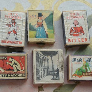 May include: A collection of vintage matchboxes with colorful illustrations and text. The boxes feature images of a brewmaster, a woman in traditional Italian dress, a man reading, a monkey, a building, and Maid Marian. The text includes "Pick Flowers", "Bitter", and "Safety Matches".