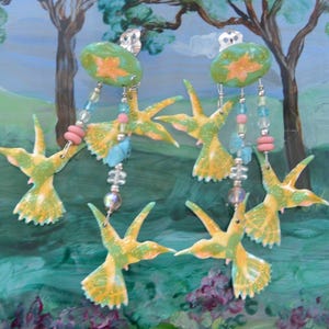 May include: Handmade earrings featuring a hummingbird design. The earrings have a green and yellow color scheme with a glittery finish. They are adorned with beads in shades of pink, blue, and clear. The background is a painted landscape with trees.