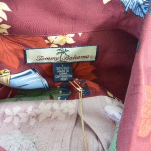 May include: Close-up of a burgundy silk shirt with a floral pattern. The shirt features a collar and a tag that reads "Tommy Bahama" and "100% Silk Made in China Copyrighted Print."
