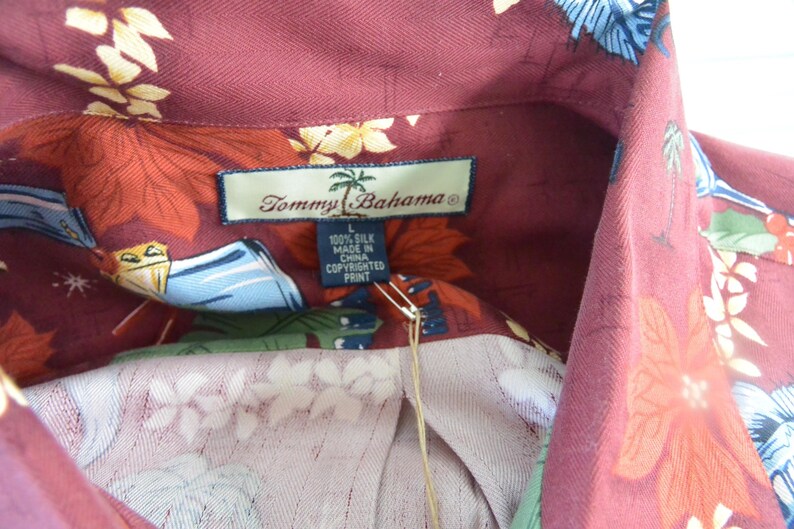 May include: Close-up of a burgundy silk shirt with a floral pattern. The shirt features a collar and a tag that reads "Tommy Bahama" and "100% Silk Made in China Copyrighted Print."