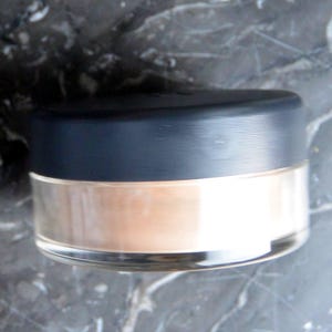 May include: A round, clear jar with a black lid containing a light brown powder. The jar is sitting on a gray, textured surface.