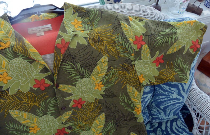 K&ouml;nnte beinhalten: Olivgr&uuml;nes Button-Down-Hemd mit tropischem Blumenmuster. Das Hemd hat gro&szlig;e gr&uuml;ne und gelbe Blumen, rote Akzente und Palmfarn. Das Hemd ist aus leichtem Stoff gefertigt und hat ein rotes Futter.