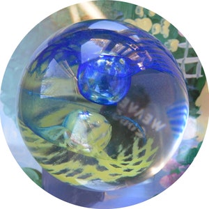 Puede incluir: Un pisapapeles de vidrio transparente con remolinos azules y amarillos y una burbuja azul en su interior. El pisapapeles está sobre una superficie con un patrón floral.