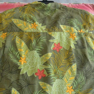 K&ouml;nnte beinhalten: Olivgr&uuml;nes Hemd mit tropischem Blumenmuster. Das Design zeigt gro&szlig;e gr&uuml;ne und gelbe Bl&auml;tter, orange und rote Bl&uuml;ten und einen detaillierten botanischen Druck. Das Hemd ist aus leichtem Stoff gefertigt.