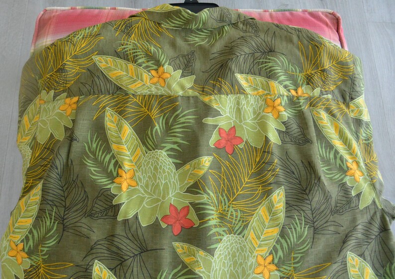 K&ouml;nnte beinhalten: Olivgr&uuml;nes Hemd mit tropischem Blumenmuster. Das Design zeigt gro&szlig;e gr&uuml;ne und gelbe Bl&auml;tter, orange und rote Bl&uuml;ten und einen detaillierten botanischen Druck. Das Hemd ist aus leichtem Stoff gefertigt.