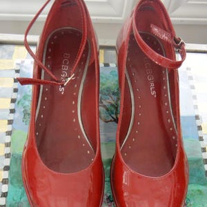 以下が含まれることがあります： 光沢のある赤いBCBGIRLSメリージェーンシューズのペア。丸いつま先、サイドバックルストラップ、小さなヒールが特徴です。靴の内側は、白いドットが入った明るい赤色です。