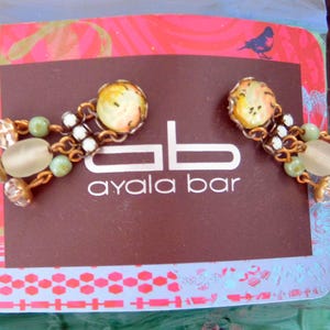 May include: A pair of Ayala Bar earrings. Each earring has a round, yellow and orange patterned stone, linked by a gold chain to a cluster of white and green beads and a larger, oval stone. The earrings are on a brown card with the text "ayala bar".