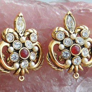 May include: A pair of ornate gold-tone earrings. Each earring features a central red stone, surrounded by clear crystals and small faux pearls. The design includes a large, clear crystal at the top and a loop at the bottom for attachment.