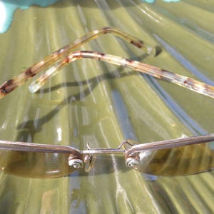 May include: Sunglasses with a silver frame and tinted lenses. The temples have a tortoiseshell pattern. The glasses are resting on a green, textured surface. The glasses have a unique design.