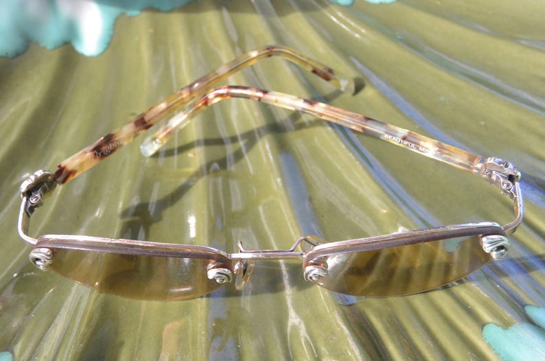 May include: Sunglasses with a silver frame and tinted lenses. The temples have a tortoiseshell pattern. The glasses are resting on a green, textured surface. The glasses have a unique design.